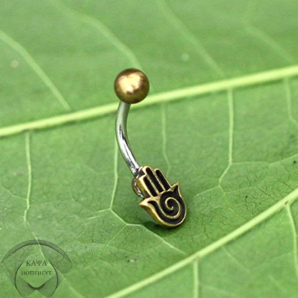 The HAMSA HAND Belly Piercing Ring in Gold by Kaya Movement, made of surgical steel, showcases a golden ball on one end and a gold-colored Hamsa hand charm with a spiral design in the center on the other. It rests elegantly on a green leaf background, making it perfect for a stylish navel piercing.